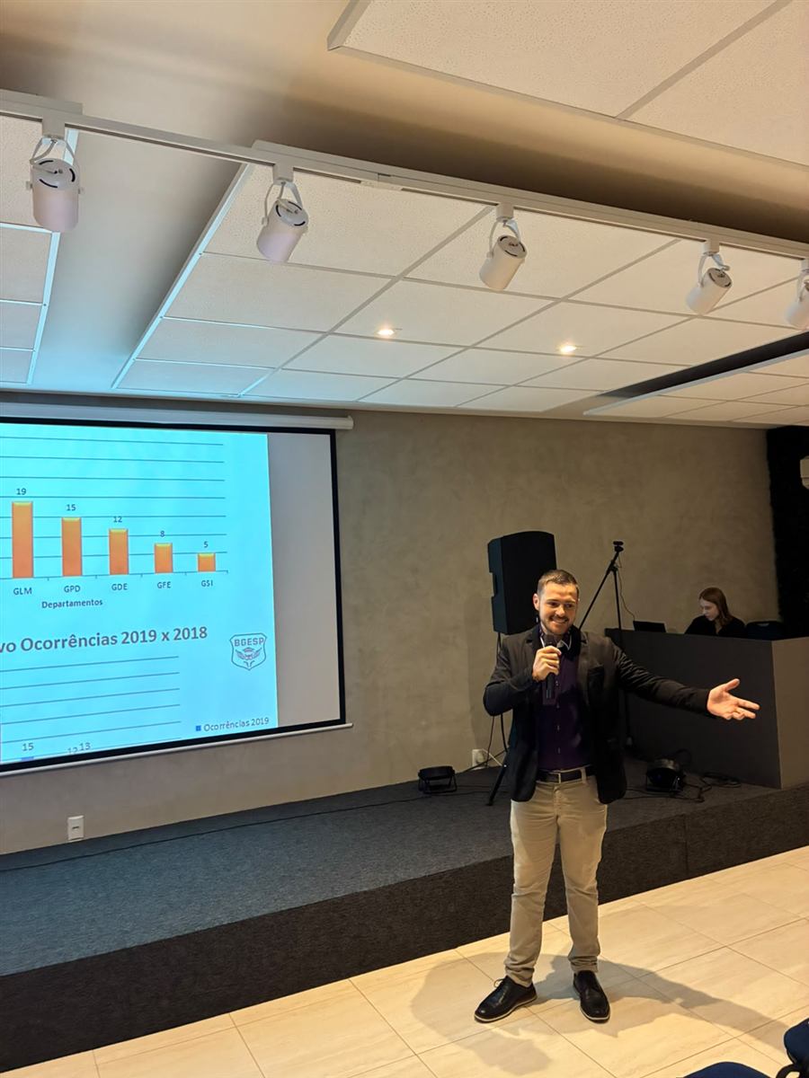 Leonardo Lauxen no palco, de braço aberto, conduzindo palestra com telão ao fundo
